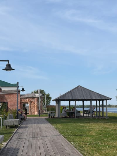 The image shows a scenic outdoor area with a wooden boardwalk leading to a covered pavilion by the water. The pavilion, constructed of weathered wood, houses several picnic tables where people are seated, suggesting a recreational or dining setting. Beyond the pavilion, a calm body of water is visible, with a treeline on the opposite bank. In the foreground, a rustic brick building with an arched opening and some partially ruined sections indicates a historical or preserved site. A wooden bench is placed along the boardwalk near the brick structure, and two individuals are interacting near it. The sky is clear and blue, with a few faint clouds. This setting is part of the museum-estate "Ustyugov House" in the city of Berezniki, Russia, which is a cultural heritage site known for its historical architecture and scenic riverfront location.