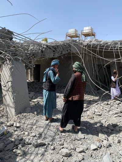 This image depicts a scene within the city of Mandisar, Afghanistan, featuring the extensive demolition or damage of a concrete building under a clear blue sky. The ground throughout the visible area is covered uniformly with a deep layer of pulverized concrete rubble, fractured debris, and sections of exposed, bent steel rebar.
The structure itself shows a collapsed roof, from which numerous twisted and tangled steel reinforcement bars protrude downwards. A large, partially intact concrete column stands upright on the left side of the frame. In the background, two elevated cuboid structures, consistent with water tanks on wooden platforms, are visible atop a distant building or elevated terrain.
Three adult males are present. On the left, a man wearing a light blue turban, light blue long shirt, and a dark vest stands facing right, his right hand raised and open in a gestural motion. He appears to be engaged in conversation with a second man, positioned centrally, who wears a green and white checkered head covering, a dark long shirt, dark shalwar pants, and a brown patterned vest. This second man stands with his back partially to the viewer, looking towards the first man. To the far right, a third man, dressed in a white long shirt and a dark vest, is partially visible, holding a slender object, possibly a stick or pipe, with both hands while observing the debris.