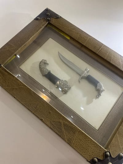 This media file displays a framed decorative dagger set, likely an ornamental piece or a historical artifact, presented against a plain, light-colored wall. The setting appears to be indoors, possibly a home or a display area, given the presentation. The frame itself is a prominent feature, constructed from a textured, leathery material in a brown hue, with decorative metal corner brackets that add a rustic and antique feel. The frame is angled, giving a dynamic perspective to the image.
Inside the frame, a dagger and its sheath are meticulously arranged on a neutral-colored background, likely matte board. The dagger is the primary object, with a polished metal blade that catches the light, indicating it's sharp. The handle is adorned with intricate silver-toned metalwork, depicting what appears to be an animal head, possibly an elephant or a mythical creature, with detailed tusks and ears. The sheath, also featuring similar elaborate metalwork, is placed below the dagger, showcasing its ornamental design. The overall presentation suggests a display of craftsmanship and cultural significance.
The lighting in the scene is bright, causing reflections on the glass covering the display and highlighting the metallic elements of the daggers. There are no people visible, and the focus is entirely on the objects within the frame. The time of day and weather are not discernible from the image. The location cues provided, "Giginyu, Nigeria," suggest that this item might be of local significance or represent a cultural artifact from that region. There is no visible text in the image.