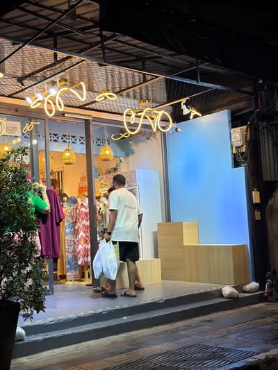 A man in a white t-shirt and black shorts is standing outside a shop, holding two plastic bags. The shop appears to be a clothing store, with garments displayed in the window. Wicker pendant lights hang inside the store. Neon signs are visible on the exterior of the building. The scene is illuminated by artificial lighting, suggesting it is nighttime. The man is facing away from the camera, towards the shop entrance. The background shows a blue wall and wooden shelving units. The setting appears to be a street in Phuket City Municipality, Thailand.