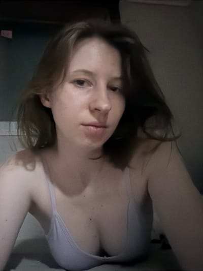 The image is a close-up, low-angle selfie of a young woman with light brown hair and fair skin. She is looking towards the camera with a neutral expression. She is wearing a light-colored tank top or bra. The background appears to be an indoor setting with muted lighting, and a dark wall or surface behind her. The image is grainy and has a slightly blurred quality. The exact location within Dongducheon-si, South Korea, is not determinable from the visual information provided in the image.