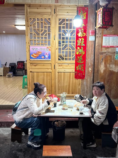 Two individuals are seated at a small rectangular table outdoors, likely at a street eatery in Gongtan, China. The scene is illuminated by a single exposed light bulb hanging centrally from a black cord.

On the left, a woman with dark hair tied in a bun and wearing glasses, a white patterned jacket, and blue jeans, is actively eating from a small bowl with a spoon. A light blue backpack rests on her left shoulder. She is seated on a small dark wooden stool.

Facing the camera on the right, another woman with short dark hair, wearing a white and black color-blocked jacket and dark pants, holds a spoon above a bowl of food. She is also seated on a small dark wooden stool.

The table between them holds several small bowls, plastic cups with lids (one containing a dark liquid), a container with chopsticks, and various small paper packets. A black cylindrical trash bin is positioned beneath the table near the left woman. An additional, empty small wooden stool is in the foreground.

The background features light-colored wooden double doors with intricate lattice-work windows. A poster advertising "Crispy Fresh Meat Pie" (香酥鲜肉饼) with a cartoon pig is affixed to the left door. To the far left, an interior space with a light wall and a few chairs is visible through an open doorway. To the right of the doors, a long red banner with gold Chinese characters hangs vertically from a wooden beam. Further right, a traditional red wooden lantern with decorative cutouts hangs from another wooden beam. The ground is a dark, uneven surface.