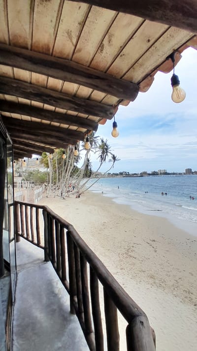 The image presents a detailed view from under a wooden veranda or balcony overlooking a sandy beach and the Caribbean Sea in Pampatar, Venezuela.
In the foreground, the structure from which the photo is taken features a rustic wooden ceiling composed of light-colored planks supported by darker, thicker wooden beams. Several vintage-style incandescent light bulbs, suspended by dark cords, hang from this ceiling. The edge of a tiled roof, with reddish-brown undulations, is visible along the top right perimeter. A dark, reflective vertical surface, possibly a window frame, is partially visible on the far left. A dark wooden railing with vertical slats and a horizontal handrail runs diagonally across the lower right portion of the image, suggesting an elevated viewpoint. The floor beneath the railing is a light gray concrete surface.
The midground reveals a wide expanse of light beige sand, forming a curved beach shoreline. Numerous slender palm trees, many leaning distinctly towards the ocean, line the back edge of the beach, providing scattered shade. Several individuals are present on the beach; some are seated or lounging near the base of the palm trees, while others are closer to the water's edge. Further down the beach to the left, dilapidated or rustic structures are visible.
The background features the calm, blue waters of the ocean, with gentle white foamy waves breaking near the shore. Multiple people are scattered in the water, swimming or wading. In the distant background, across the bay, a low-rise urban landscape with various buildings and structures is visible under a slightly hazy sky. The sky above is a light blue with scattered white, fluffy clouds. The overall lighting suggests a bright daytime scene.