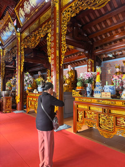 A woman with dark hair is praying in what appears to be a Buddhist temple. She is wearing a black shirt and rose-colored pants. A small purse is visible across her shoulder. The temple's interior features red carpets and elaborately decorated wooden structures, with gold accents. In the background, there are statues and offering tables with flowers and other religious items. The temple has columns covered in gold lettering. The location is Diên Khánh, Vietnam.