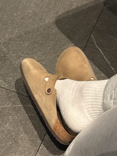 The image shows a person wearing a pair of beige suede Birkenstock Boston clogs with white socks. The person is standing on a gray tile floor. The exact location within Heroica Puebla de Zaragoza, Mexico, cannot be determined from the image.