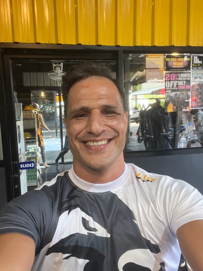 This is a selfie of a man with short dark hair and a beaming smile, wearing a white and black athletic shirt. He appears to be in a gym or fitness center, as indicated by the posters and displays visible in the background behind him. The setting features a yellow corrugated metal wall on the left and a large glass window or display case on the right. Through the glass, signs promoting gym memberships and class schedules are visible, including "Day Pass - Gym Bowl 900 Baht", "20% OFF 1 MONTH OPEN GYM PASS 2000 THB", and "GROUP CLASSES" with specific times like "10.30 AM", "11.45 AM", and "19.00PM". A mannequin is also displayed behind the glass. The overall impression is a cheerful, energetic atmosphere, suggesting the man is enjoying his time at the facility. The presence of "Baht" indicates the location is Thailand.