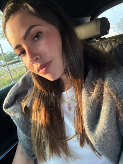 A selfie captured from a low angle shows a woman with brown hair wearing a gray cardigan over a white shirt. A necklace with a turtle charm is visible. She is inside a car, judging by the car seat headrest in the background. Through the car window, a blurred landscape with greenery and a few buildings can be seen. The location is Tlaxcalancingo, Mexico.