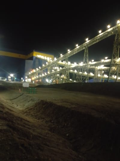 The image depicts an industrial site at night in Қопа ауылдық округі, Kazakhstan. A large conveyor belt system, illuminated by numerous lights, stretches across the frame, supported by a network of metal structures. To the left, a substantial blue and yellow building is partially visible. The foreground is a rough, unpaved area with mounds of dirt. The overall scene suggests ongoing industrial activity in a rural or semi-industrial setting.