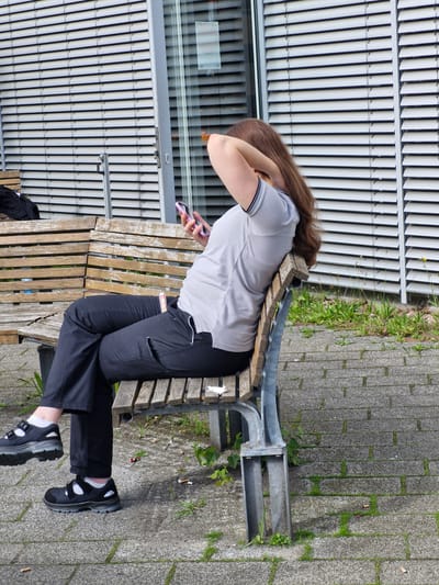 A woman with long brown hair is sitting on a slatted wooden bench outdoors, holding a pink smartphone in her right hand and resting her left hand on the back of her head. She is wearing a light gray polo shirt with dark trim on the collar and sleeves, and black pants. Her black shoes have white socks showing. The bench is situated on a paved area with gray interlocking bricks, and there is some sparse green vegetation growing between the bricks. Behind the bench is a modern building with large, horizontal white blinds covering a glass facade. The scene appears to be a public space in the city of Bremen, Germany, possibly near a commercial or administrative building given the architecture. There are several discarded cigarette butts on the ground around the bench, suggesting it may be a designated smoking area.