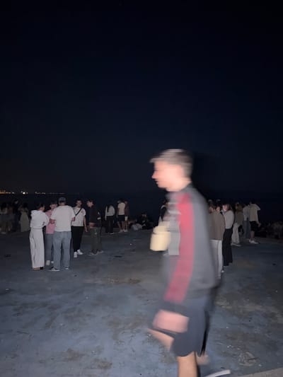 At night in Grad Rijeka, Croatia, a crowd of people is gathered on a dark, concrete surface, possibly a rooftop or pier. Many are wearing light-colored clothing, including white shirts and pants. There is a blurred figure wearing a gray and red jacket and dark shorts in the foreground. The background includes a dark night sky and what appears to be the city lights of Grad Rijeka in the distance.