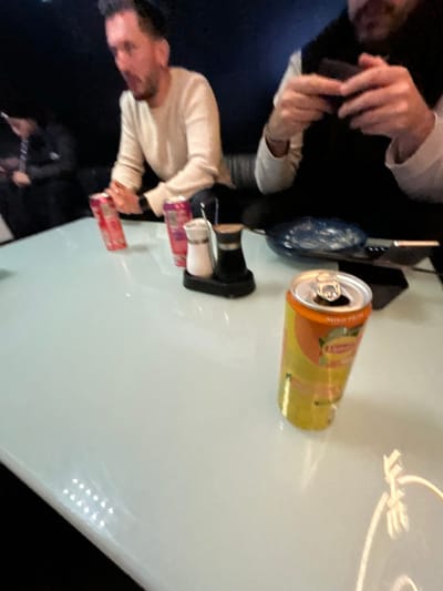 The image depicts an indoor scene at what appears to be a restaurant or bar in Illkirch-Graffenstaden, France, focused on a light-colored, reflective table surface. Three individuals are partially visible in the low-lit background.
On the left, a male subject with dark hair and a beard, wearing a light-colored long-sleeve shirt, is seated, with his mouth slightly open, suggesting he is speaking or reacting. Two red or pink beverage cans, one appearing to be a Red Bull, are situated near his position on the table.
On the right, another male subject, wearing a dark vest over a light long-sleeve shirt, is seen holding a dark rectangular object, likely a smartphone, with both hands, indicating interaction with the device.
In the immediate foreground, on the right side of the table, a yellow Lipton Iced Tea (Peach flavor) can, with an open tab, stands upright. Further back on the table, centrally located, is a black caddy holding white salt and dark pepper shakers. A dark blue or patterned plate or bowl, and a dark rectangular object (possibly another phone or menu) are also visible on the table. A metal utensil handle is partially visible near the plate. A third individual is vaguely discernible in the far left background, also seated. The environment features dark upholstered seating and artificial lighting, creating reflections on the table surface.