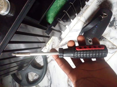 A close-up, overhead shot shows a hand holding a screwdriver with a red and black handle. The screwdriver is positioned horizontally, with its metal shaft extending to the left. Behind the screwdriver, a black steering wheel is visible, partially obscured by the hand and tool. In the background, a black speaker with a fabric texture and a strap is lying on a marbled tile floor. The overall scene appears to be indoors, possibly in a domestic setting in Keffi, Nigeria.