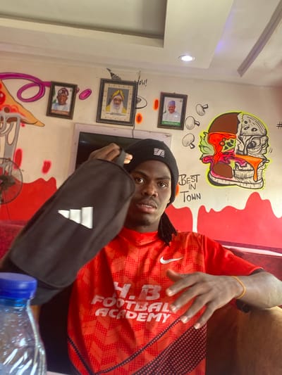 A man is indoors, potentially in a restaurant in Tukur Tukur, Nigeria. He wears a black beanie and an orange athletic jersey. He holds a black Adidas bag. Behind him, the wall is decorated with a cartoon burger, framed portraits, and graffiti. A water bottle sits on the table in the foreground.