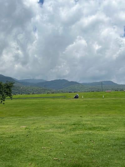 The image depicts a vast, rolling green landscape under a cloudy sky, reminiscent of a park or recreational area. In the middle ground, a golf cart is visible on a pathway that runs horizontally across a manicured lawn. Beyond this pathway, several benches are spaced out, suggesting a public seating area. Further in the background, a dense, tree-covered mountain range stretches across the horizon, shrouded in mist, indicating a natural, possibly tropical environment. The foreground features more lush green grass, with some scattered leaves and debris. The overall impression is one of a serene, expansive outdoor space with a blend of cultivated and natural elements. The specific location within Chiang Mai City Municipality, Thailand, is not explicitly evident from the visual cues alone, but the presence of lush greenery and misty mountains suggests a location with such geographical features.