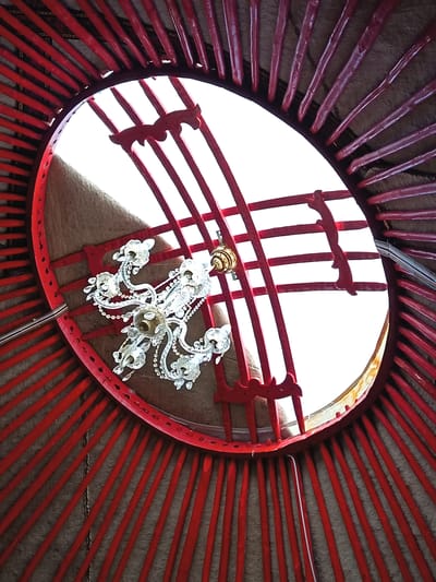 A low-angle interior view looking up at the crown of a yurt in Tosor, Kyrgyzstan, reveals a circular opening against a bright sky. The ceiling structure is made of red wooden poles radiating outwards, connected to a circular frame at the top. Within the opening, a decorative chandelier with crystal elements hangs from the wooden lattice. The interior walls of the yurt are covered with beige-colored fabric, and the red poles appear to be attached to this fabric. Two red decorative wooden cutouts, resembling stylized animal heads or horns, are attached to the lattice structure above the chandelier. A white wire runs vertically along one of the red poles on the right side of the frame. The lighting is uneven, with the bright sky creating blown-out highlights around the opening and casting shadows on the interior elements.