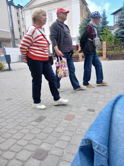 Three individuals are walking on a paved sidewalk in Truskavets, Ukraine. The person on the left is a woman with blonde hair, wearing a red and white horizontally striped long-sleeved shirt and dark blue pants. She has a dark red purse slung across her body. In the middle is a man wearing a dark gray t-shirt, a dark jacket with maroon and black accents, blue jeans, and a blue and white polka dot hat. He is carrying a multi-colored shopping bag. The person on the right, also a man, is wearing a gray shirt, blue jeans, and a dark jacket. He has a light-colored crossbody bag. Behind them, there is a building with light-colored facade and dark windows, trees, and wooden benches. The sidewalk is made of grey paving stones.