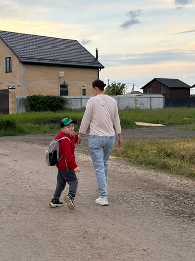 The image depicts a woman and a boy walking on a dirt road in what appears to be a residential area. The boy is wearing a red jacket, gray pants, black and gold sneakers, a green baseball cap, and a gray backpack. The woman is wearing a long-sleeved white shirt, blue jeans, and white sneakers. The boy is holding a white object in his right hand. The location is in Rossiyskiy, Russia. There are houses and green vegetation along the roadside. The sky is visible with some clouds.