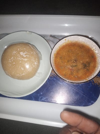 On a serving tray with a marble-like pattern, sits a plate of what looks like wrapped dough, possibly fufu or similar starchy staple food. Beside it is a bowl of reddish-brown soup or stew, seemingly containing some herbs and spices, also wrapped in thin film. A human finger is partially visible on the tray's edge. The food presentation suggests a meal ready to be consumed.
