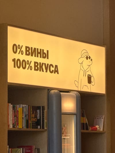 The image depicts a brightly lit sign with the text "0% ВИНЫ 100% ВКУСА" (0% WINE 100% TASTE) in large brown letters. To the right of the text, a stylized black and white illustration of a dog wearing a beanie and sunglasses, and holding a mug, is present. The sign is mounted above a bookshelf filled with various books. To the right of the bookshelf is a refrigerator or display case with a lit interior. The overall environment appears to be an indoor establishment, possibly a cafe or shop. The city of Irkutsk, Russia is not visually identifiable within this image.