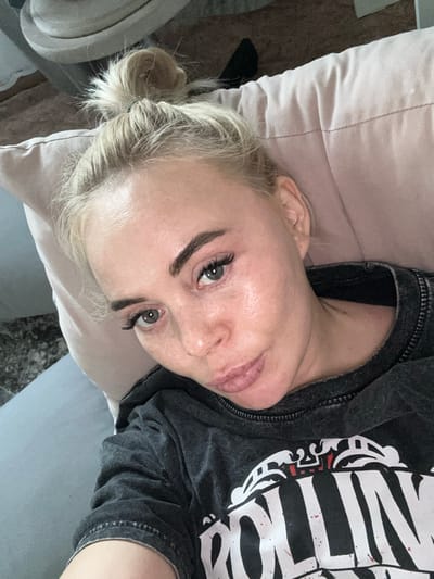 The image shows a fair-skinned person with blonde hair pulled back in a bun, lying on a light pink pillow and a gray sofa. The person is wearing a dark gray t-shirt with a white and black graphic print. The person's face is turned towards the camera. The location is unknown and there is no evidence of the city of Высокий, Russia.