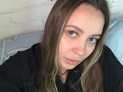 A selfie depicts a woman with light skin, light eyes, and light brown hair with blonde highlights. She has a silver nose ring. She is wearing a black shirt. The background appears to include a white brick wall on the bottom and a blue patterned surface on top. There is no clear evidence in the image to suggest this photo was taken in Novopokrovskaya, Russia.