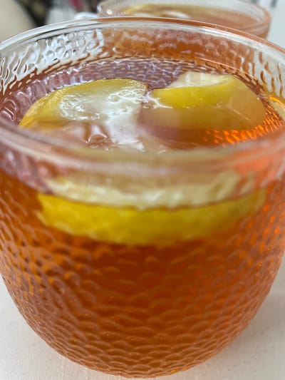 A close-up shot focuses on a clear glass filled with a reddish-orange liquid, likely iced tea or a similar beverage. Two thick slices of lemon are visible submerged in the liquid, with one slice resting near the top and another further down. The glass has a textured, patterned surface, creating an irregular, dimpled effect on the exterior. The background is out of focus, showing hints of another glass and a light-colored surface. The lighting appears natural, casting reflections on the glass and liquid. The image is shot from a slightly elevated angle, looking down into the glass. There is no identifiable location information, and no people or animals are present.