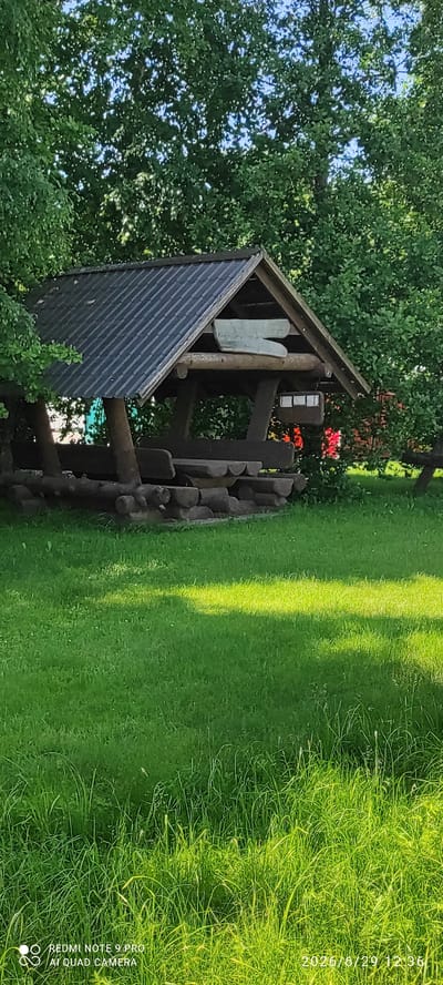 A wooden gazebo with a dark, sloped roof stands in a grassy area surrounded by lush green trees. The gazebo features rustic wooden benches and a table made from logs. A light-colored, abstract wooden sculpture is mounted on the upper part of the gazebo's front. In the background, hints of red and green are visible amongst the foliage. The foreground is dominated by a well-maintained lawn, with patches of sunlight creating a dappled effect. The timestamp "2026/6/29 12:36" and "REDMI NOTE 9 PRO AI QUAD CAMERA" are visible in the lower right and left corners, respectively.