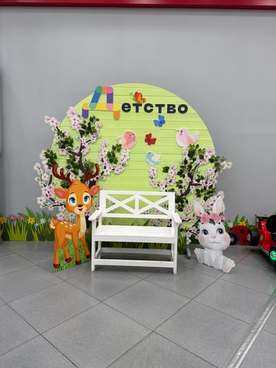 This media file depicts a colorful, whimsical cutout display likely intended for children, possibly in a play area, mall, or event space in Khabarovsk, Russia. The central subject is a large, light green, semi-circular backdrop with vertical wooden planks. On this backdrop, a stylized cherry blossom tree with pink flowers and green leaves is depicted.

Various cartoon characters and elements are placed around the tree. A white rabbit with pink accents on its ears and paws, adorned with a floral crown, is positioned at the top right, appearing cheerful. Below and to the left of the rabbit, a light pink bird with a darker pink head is perched on a branch. To the left of the tree, there are colorful, abstract shapes resembling building blocks, and further down, two butterflies (one blue, one red) are depicted. A light blue bird is also visible near the red butterfly.

At the bottom center, a fawn with large blue eyes and brown antlers is featured, looking curiously towards the viewer. It is situated in front of a white, decorative fence-like structure. The background behind the characters and tree consists of green grass and small flowers, suggesting an outdoor meadow or garden scene.

Partially visible at the very top of the image is a red toy vehicle with a ladybug design, emitting green lights. Visible text on the green backdrop, running vertically along the left side, reads "ДЕТСТВО" (childhood) in dark, bold letters. The flooring appears to be grey tiles laid in a diamond pattern, a common sight in indoor public spaces. The lighting suggests it is daytime, and there are no indicators of specific weather conditions. The overall mood conveyed is playful and inviting for children.