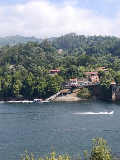 This image depicts a scenic view of the Cávado River in Terras de Bouro, Portugal. The river, with its dark blue, rippling water, dominates the foreground. In the middle ground, a tree-covered hillside rises steeply from the riverbank. Several buildings, including houses and shops, are nestled into the slope of the hill, some with red-tiled roofs. A bridge with stone arches spans the river on the right side of the image.
On the river, a white passenger boat with "VIEIRA MINHO" written on its side is docked at a pier with a ramp leading to the shore. Further out on the water, a jet ski with two people on board is creating a white wake as it speeds across the surface. The background features more hills covered in dense green trees, with a few power lines visible traversing the landscape. The sky above is a mix of blue and white clouds.