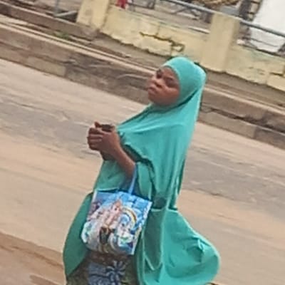 This tilted, somewhat blurry daytime image captures a woman standing or walking outdoors, possibly in Dadi Riba, Nigeria. The woman, who appears to be of African descent, is dressed in a teal-colored head covering (hijab) and a matching, loose-fitting outer garment that drapes over her body. Her hands are clasped in front of her, and she carries a small light blue shoulder bag adorned with a colorful, illustrative design featuring what appears to be a unicorn or fantasy creatures among clouds. A patterned fabric, possibly a skirt or dress, is visible beneath her outer garment. Her facial expression is neutral or contemplative. The background consists of a worn, light-colored building wall or structure, showing some texture and what may be dirt streaks, and a rustic, unpaved ground surface. There is no discernible text or specific activity beyond her presence in the outdoor setting.