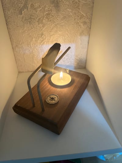A close-up, angled shot shows a wooden object with a lit tea light candle and a coin on top. The wooden object is rectangular with a beveled edge and a light brown finish. A metal stand, possibly for incense, is attached to the object, holding a piece of wood that is partially burned. A small, round coin with intricate markings is placed on the wooden surface to the left of the candle. The background consists of a textured white wall and a white shelf. The lighting is dim, with the candle flame casting a warm glow. The scene appears to be indoors, possibly on a shelf or table. There are no people visible in the image. There is no information that can be used to determine the location in the city of Krasnyye Barrikady, Russia.