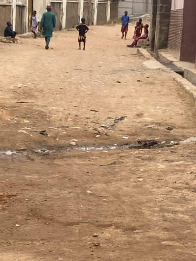 The image depicts an outdoor scene in Minna, Nigeria. Several people are present, including individuals wearing what appears to be a traditional green garment, a blue garment, and what appears to be a uniform. The ground is a tan-colored, uneven surface. The backdrop includes partial views of buildings, possibly constructed with brick, suggesting the scene is located in an urban or semi-urban environment. The primary activity observed appears to be walking or standing.