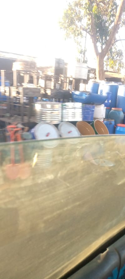 An outdoor storage area appears with an assortment of containers. There are metal drums and bins, some with white or brown lids, along with stacks of blue plastic barrels. A tree with sparse foliage is in the background. The image seems to be taken from inside a vehicle with a dirty windshield, causing a blurry foreground.