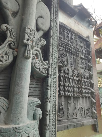 The image depicts intricately carved stone or concrete structures, likely part of a temple or religious site. The structure on the left features stylized floral and animal motifs, with a weathered texture. The structure on the right showcases a series of carved figures and geometric patterns, arranged in distinct registers or panels. The overall scene suggests a site with cultural and historical significance, located in Muang Pattaya, Thailand. The style of the carvings is reminiscent of Southeast Asian religious art. The architectural design features decorative details and potentially represents elements of local folklore or religious narrative.
