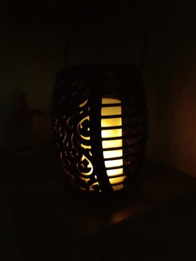 An ornate, dark-colored lantern with a curved, basket-like design is emitting a warm, yellow light from its interior. The lantern has decorative cutouts that allow the light to shine through, creating a patterned effect. The lantern is placed on a dark surface, likely a table or shelf, in a dimly lit environment. The background is dark and indistinct, with a faint shadow of a person or object visible on the left side, suggesting an indoor setting. The overall impression is one of atmospheric and decorative lighting.