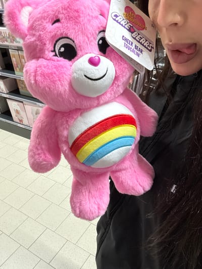 A pink Care Bear plush toy, specifically "Cheer Bear" with its characteristic rainbow belly badge, is held in the foreground. The toy displays its original retail tag, which includes "Les Bisounours," "Care Bears," "Cheer Bear," and "Toucalin." A person with long, dark hair and wearing a black jacket is partially visible in the upper right quadrant, holding the toy. The person's nose, mouth, and chin are visible, with the tongue partially extended from the mouth. The scene is an indoor retail environment. In the background, light-colored metal shelving units display various boxed merchandise, some partially obscured. The floor is covered with light-toned square tiles. The lighting is uniform artificial illumination. The precise retail establishment within Sint-Niklaas, Belgium, cannot be determined from the visual information provided.