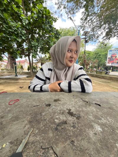 A young Indonesian woman wearing a light gray hijab and a striped sweater is seated at a rough concrete table in an outdoor park setting. She is looking to her right with a slight smile. Her arms are crossed on the table in front of her. In the foreground of the table, there is a red elastic band and a key. The background shows lush green trees, a street, a billboard, and what appears to be a building with red roofs in the distance. The park appears to be a public space in the city of Langsa, Indonesia, possibly a recreational area with outdoor exercise equipment visible to the left of the woman. The sky is partly cloudy.