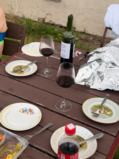 The image shows a picnic scene on a wooden table. Several objects are present, including a bottle of red wine with two glasses partially filled, two decorative plates with illustrations, a fork and spoon, a bottle of Coca-Cola, and a clear plastic container with food. A crumpled piece of aluminum foil is also visible. The setting appears to be outdoors, with grass and concrete visible in the background, suggesting a casual gathering. The lighting and shadows indicate it is daytime, likely afternoon, with pleasant weather. One person's legs, wearing blue and green swim trunks, are visible in the lower left corner, hinting at a recreational activity, possibly near water given the attire. There are no discernible emotions expressed, but the arrangement of items suggests a meal or gathering is underway. Some text is visible on the wine bottle label and on the Coca-Cola bottle, but it is not legible enough to be transcribed. The overall scene depicts a relaxed outdoor meal.