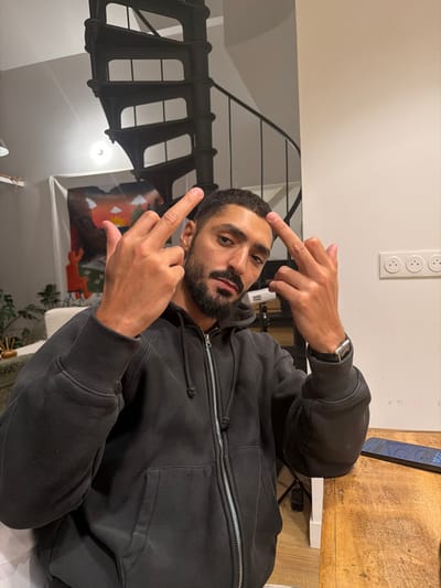 The image shows an adult male seated at a wooden table indoors, within a residence located in Saint-Denis, France. The subject, who has short dark hair and a full beard, wears a dark gray or black zippered hooded sweatshirt and a silver-faced watch with a black strap on his left wrist. He is looking directly at the camera, simultaneously extending the middle finger of each hand upwards.

In the background, a prominent black spiral staircase ascends on the left. Below the staircase, a white wall displays a tapestry or artwork depicting an orange sun, a landscape, and stylized red figures. A wall-mounted light fixture is visible near the top left. To the right of the subject, a plain white wall features a triple electrical outlet. On the wooden table to the subject's right, a black smartphone displaying the time "22:14" is visible.