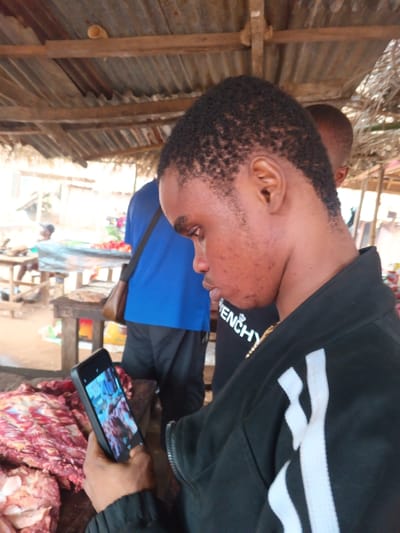 The image depicts an outdoor market scene in Omicha Umu Awada, Nigeria. In the foreground, a young dark-complexioned male with short, dark, textured hair is observed in profile, looking down at a black smartphone held horizontally in both hands. He wears a black zippered jacket featuring two white vertical stripes on the right sleeve and a black t-shirt with a white "GIVENCHY" logo visible beneath the jacket. A gold chain is around his neck. His face exhibits visible acne or blemishes on the chin and cheek area. The smartphone screen displays what appears to be an image of raw meat, consistent with the substantial pile of red meat visible on a wooden stall surface immediately to his left.
In the mid-ground, behind the primary subject, another male figure is partially visible, wearing a blue t-shirt and dark trousers, with a brown leather-like shoulder bag hanging from his left side. The background is blurred but shows additional market stalls constructed from rough wooden frames and corrugated metal roofs. Other individuals and market goods, possibly produce, are discernible further back. The ground appears to be dirt or earth. Overhead, the stall roof structure includes wooden beams and corrugated metal sheets, with a light-colored pipe-like object resting on one of the beams. The scene is brightly lit by natural light.