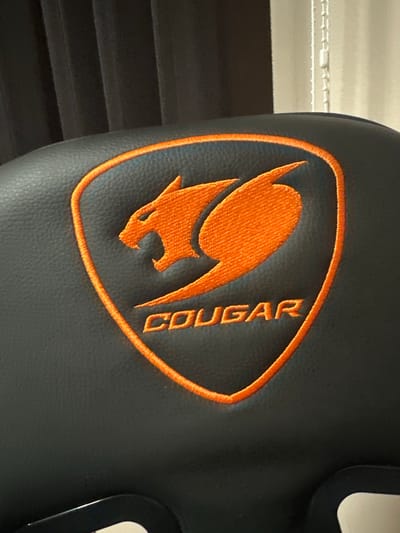 A close-up photograph captures a bright orange embroidered logo on a dark, textured surface, consistent with the backrest or headrest of an upholstered chair. The logo features a stylized cougar head facing right, with an open mouth, enclosed within a shield-shaped outline, all rendered in orange thread. Below the cougar design, the word "COUGAR" is embroidered in capital letters, also in orange. The base material is a dark grey or black synthetic leather, exhibiting a fine, pebbled texture across its surface.
In the blurred background to the left, vertical pleats or folds of a dark fabric, possibly curtains or drapes, are visible. To the right, the background is lighter, showing faint vertical lines and a distinct white beaded pull chain, characteristic of window blinds, with a small white plastic connector along its length. A small section of a dark, rigid component, possibly plastic or metal from the chair's frame or armrest, is visible at the very bottom edge of the image. The scene is illuminated by soft, ambient light. This scene is situated in Saraia, Bulgaria.