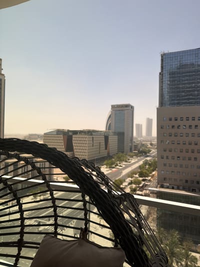 The original image is oriented vertically, appearing rotated 90 degrees counter-clockwise. Describing the scene as if correctly oriented, it presents an expansive urban view from a high vantage point, likely a balcony or terrace, in Lusail, Qatar.
In the foreground, at the bottom right, a segment of a dark, woven outdoor chair or seating arrangement, possibly made of wicker or rattan, is prominently visible. A light-colored cushion rests within the chair, suggesting a comfortable seating area.
Beyond the balcony, a modern cityscape unfolds under a bright, hazy sky. Numerous tall buildings dominate the middle and background. To the left, a sleek, reflective glass skyscraper stands out. To its right, a multi-story building with a light-colored facade features many rectangular windows. Further into the midground, a prominent building clearly displays the word "HILTON" on its rooftop sign, identifying it as a Hilton hotel. Other architectural structures, including buildings with distinctive patterned facades, extend into the distance, appearing increasingly indistinct due to atmospheric haze.
Below the cluster of buildings, a wide, multi-lane road or highway stretches through the urban landscape. It is bordered by lush green trees, including several recognizable palm trees, adding patches of greenery to the concrete environment. The sky, visible above the buildings, is a pale, uniform hue, indicating either strong daylight diffused by atmospheric dust or a very bright, overcast condition. No people are visible, and the scene captures a static moment of the bustling city. The hazy conditions suggest either a dusty day or very bright sunshine.