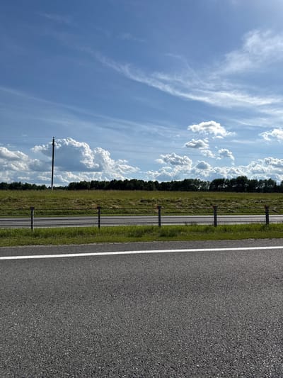 The image depicts a roadside scene, rotated 90 degrees clockwise. The foreground is dominated by a section of asphalt road with a white line marking its edge. To the left of the road, there is a grassy verge, and further to the left, a low fence with vertical posts topped with red reflectors. Beyond the fence, a line of trees forms a dark green border. The background features a vast blue sky filled with scattered white clouds of various sizes and shapes, some appearing wispy and others more cumulus-like. The overall impression is of a clear, bright day. The scene evokes a sense of open space and the calm of a rural or semi-rural environment. There are no visible people or animals. The time of day appears to be daytime, likely late morning or afternoon given the brightness of the sky. The location context provided is Пяршайскі сельскі Савет, Belarus. There is no visible text in the image.