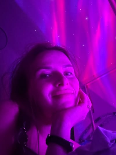 A young woman is captured in a close-up, selfie-style shot against a backdrop of vibrant pink and purple lighting that resembles an aurora borealis. She rests her chin on her hand, looking directly at the camera with a slight smile. Her hair is dark and falls around her face. The lighting casts a strong purple hue over her skin. She is wearing a black watch on her left wrist, and a necklace is visible around her neck. The environment appears to be indoors, with the lighting projected onto a wall or ceiling. The image does not provide any specific indicators of the location being Rusalevka, Russia.