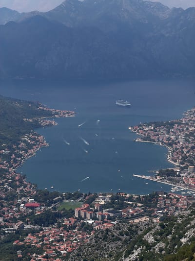 This is an aerial, high-angle, wide shot of a body of water with a city built along its shore and mountains in the background. The water is a deep blue and dotted with numerous boats, including a large cruise ship on the left side of the image. The city, appearing to be a densely populated coastal town, is characterized by buildings with red tile roofs, interspersed with greenery. There are also visible structures like a sports field. The terrain rises steeply from the water, with lush vegetation on the lower slopes and rocky, barren mountains dominating the upper background. The lighting suggests it is daytime, possibly midday, with clear weather conditions allowing for excellent visibility. The scene depicts a serene, picturesque landscape at Mirac, Montenegro, showcasing the interplay between urban development, natural topography, and maritime activity. No visible text is present in the image.