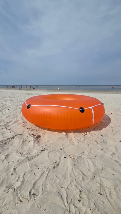 The image depicts an orange inflatable swim ring lying on a sandy beach. The swim ring is in the foreground, occupying the lower half of the frame. It is round with a hole in the center and has white rope woven through black loops attached to its sides. The sand is light in color and shows some debris like dry leaves and twigs, as well as footprints.
In the background, the sea stretches to the horizon under a cloudy sky. Several people are visible in the water, some wading and others swimming. The scene appears to be a public beach in Jūrmala, Latvia, given the presence of people enjoying the water on a summer day. The overall atmosphere is one of leisure and recreation at the seaside.