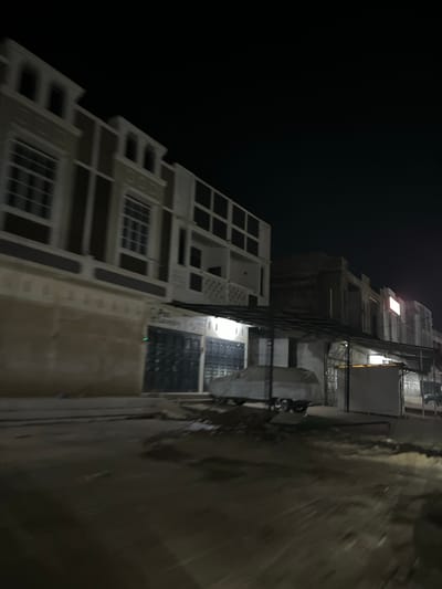 The image depicts a vertical, low-angle view of an apartment building at night in Benuaraja, Indonesia. The building appears to be under construction or renovation, with exposed concrete and scaffolding visible in some sections. The overall lighting is dim, illuminated by scattered artificial lights from within the building and the street.
The building's facade shows a mix of finished and unfinished elements. The lower sections feature pre-fabricated panels with a grid-like pattern and windows. Higher floors are more skeletal, with concrete slabs and visible structural beams. One window on an upper floor is brightly lit with an orange-ish glow, possibly indicating an occupied unit or construction activity. The street in front of the building is wet, suggesting recent rain or ongoing watering, and reflects the building's lights.
The scene conveys a sense of a developing urban environment, characteristic of a place experiencing growth. The time of day is clearly night, emphasized by the dark sky and artificial lighting. There are no people or specific activities clearly depicted, but the presence of lights suggests some level of ongoing work or habitation. The atmosphere is somewhat stark and functional due to the construction elements and the night setting. No legible text is visible in the image.