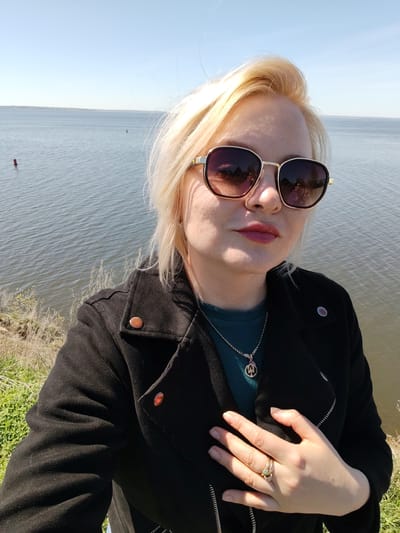 A woman is seen in a selfie outdoors in the city of Икское Устье, Russia. She has blond hair and is wearing sunglasses and a black jacket over a green top. A necklace and rings are visible. The background shows a body of water under a clear sky. Some greenery is visible at the bottom of the frame.