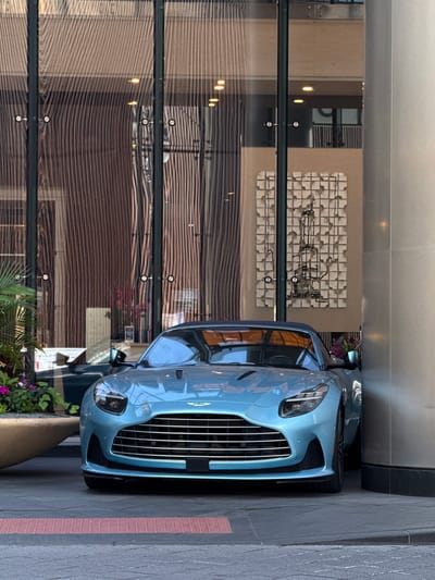 A bright blue Aston Martin sports car is parked on a sidewalk in front of a modern building with large glass windows. The car is the primary subject, with its sleek design and metallic paint reflecting the surrounding environment. The building's facade features horizontal wooden panels and a decorative wall sculpture with metallic elements.
The scene appears to be outdoors, with natural light suggesting it is daytime. The weather seems fair, as there are no visible signs of rain or harsh sun. The location is identified as Chicago, USA, though specific landmarks are not apparent. The image captures a sense of urban elegance and luxury, with the high-end car parked in a sophisticated setting.
The reflection in the glass windows shows interior lighting and architectural details of the building, creating a layered visual effect. The sidewalk is paved with textured gray stones, and a red-patterned strip runs along the edge. Small potted plants with purple flowers are visible near the car, adding a touch of color and life to the scene. No people are visible in the image. The overall impression is one of quiet sophistication, showcasing the car as a piece of art in an urban landscape.