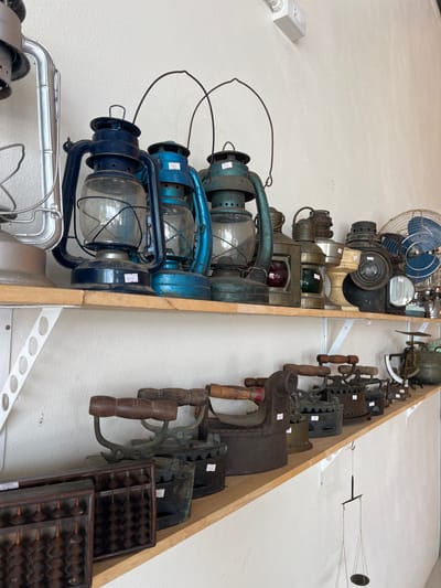 This image shows a collection of vintage items displayed on a white wall. The items are arranged on wooden shelves. On the left side, there are several old lanterns, some blue and some metallic. One blue lantern has a price tag with the number "700" visible. Below the lanterns, a shiny metallic object, possibly part of an old fan or appliance, is visible.

On the right side, a row of antique flat irons is displayed, each with wooden handles. Below the flat irons, two abacuses are stacked, both made of dark wood. The top abacus has a price tag with "1,500" partially visible.

In the background, at the top left, a vintage blue electric fan is mounted on the wall. Above the flat irons, there is a scale with a circular weight. The scene appears to be an indoor display, possibly a shop or a museum, showcasing historical tools and devices. The lighting suggests it is daytime. There are no people visible in the image. The overall impression is one of a curated collection of antique objects.